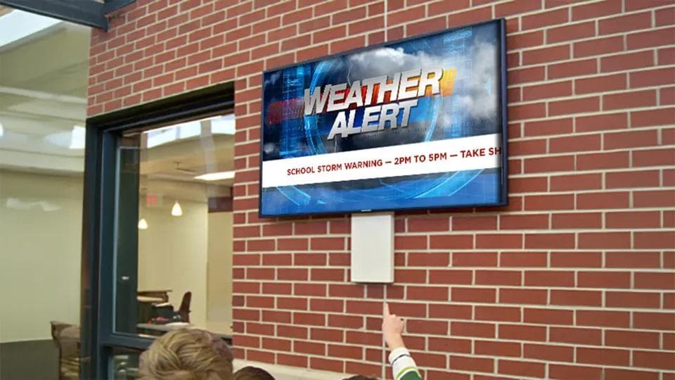 Digital signage on a brick wall in a school plays a weather alert graphic for students in the hall.