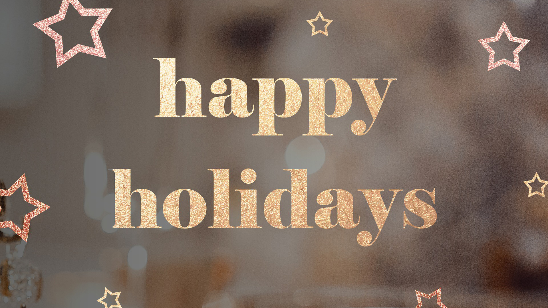 Shimmery Gold Happy Holidays Graphic with a blurred brown and gray background with small and large stars all around.