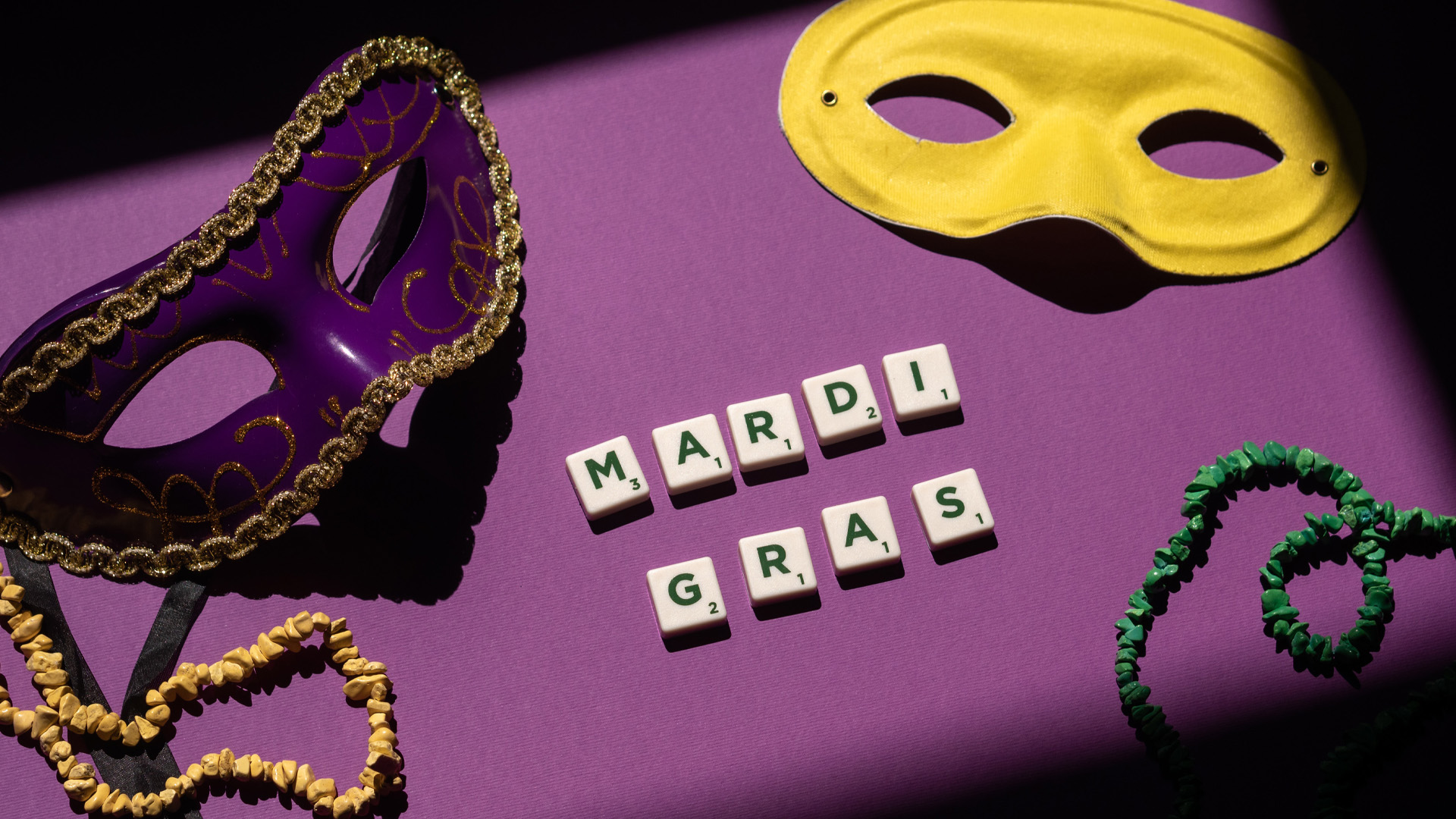 Purple background, with green and yellow stoned necklacesspread across the bottom of the graphic. Purple and yellow mask are spread across the top of the image. Scramble letters spelling our "Mardi Gras" in the center of the graphic.