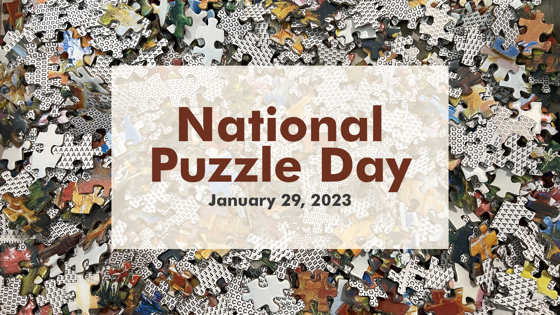 There is a photo of puzzle pieces overlapping each other laying on a table. In the middle of the graphic there's a deep red text that reads, "National Puzzle Day January 29, 2023".