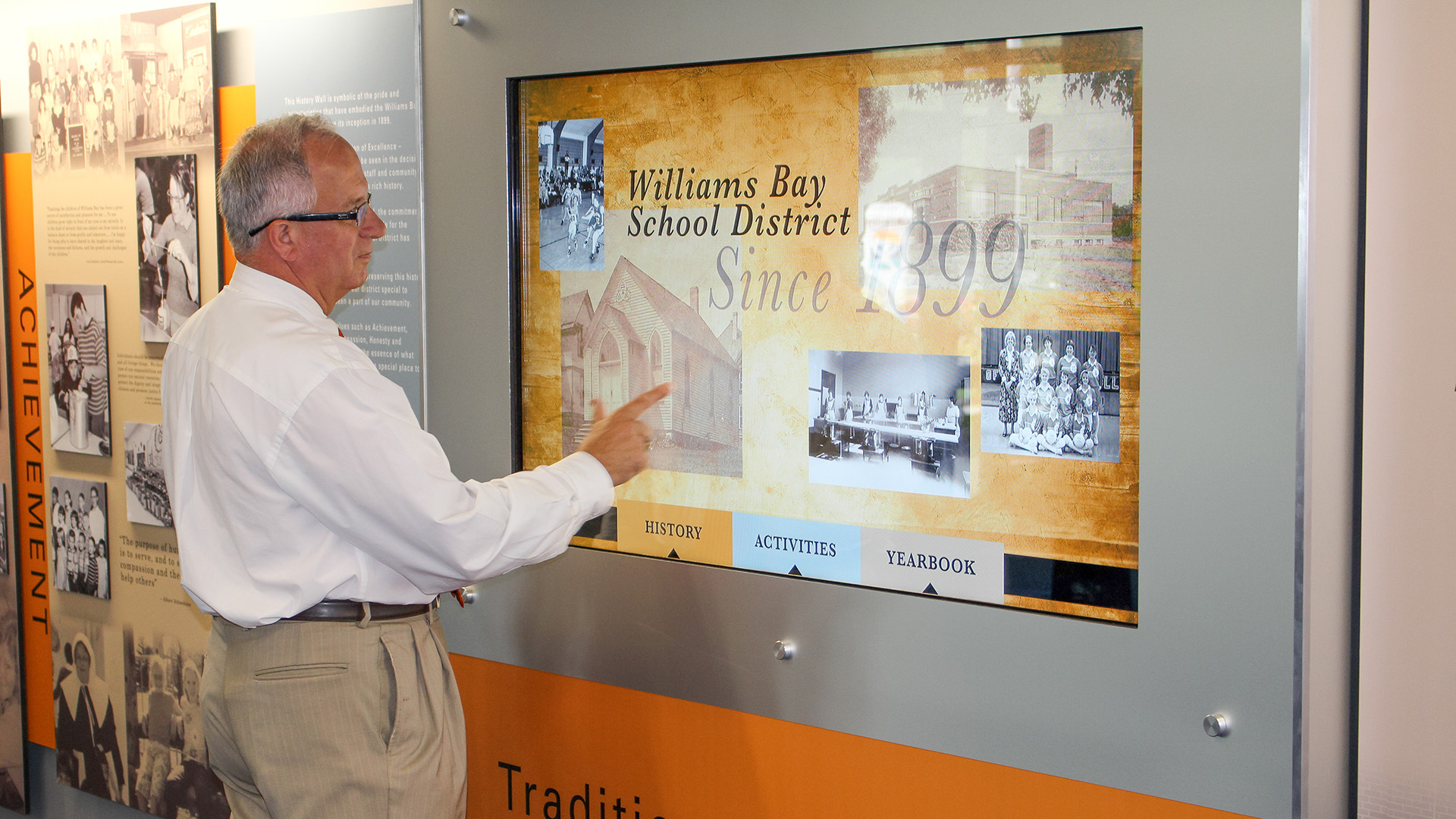 A display wall features history panels of the school in the blues and golds. A man standing at the wall is interacting with the school digital signage touchscreen display showing history and yearbook content.