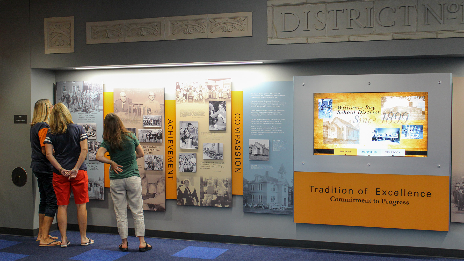 A display wall features history panels of the school in the blues and golds as students and visitors stand to read the content. Also on this wall is a school digital signage touch screen interactive display showing history and yearbook content.