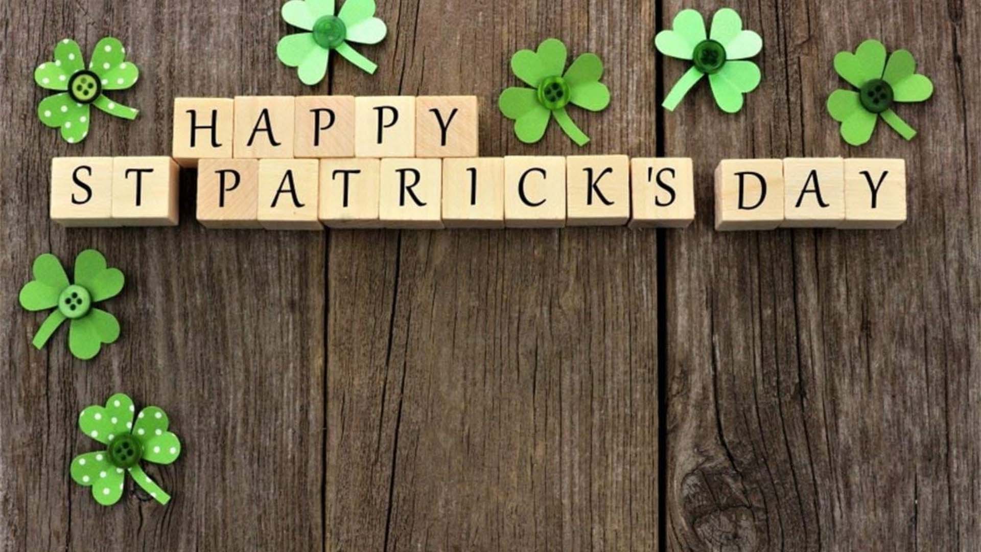 Wooded background with lighter toned wooden block spelling out Happy St Patrick's Day. Seven cut out paper shamrock aligning the left and top of the graphic with buttons in the middle of the shamrock