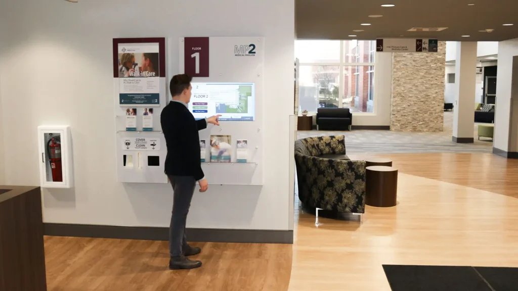 Inside the loby of a medical building with elevators and chairs. A man is standing and using a wall kiosks touchscreen display for wayfinding.
