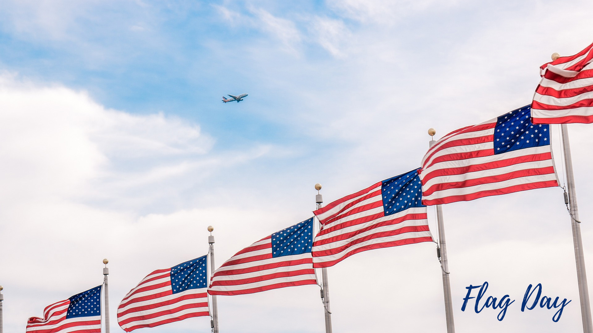 Photo of 6 flag poles with 6 USA flags waving in the wind! There is a blue sky with little cloud coverage. Toward the center of the graphic there is a airplane in the distance. In the bottom left corner in a cursive navy text it reads Flag Day.