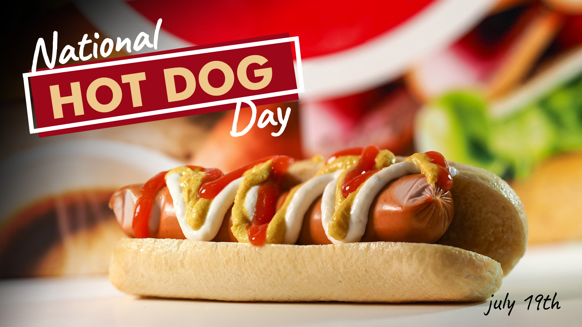 A photo of a hot dog in a bun laying on a white table with a blurred out colorful background. On the top left side of the image there is text that reads National Hot Dog Day with emphasis on the Hot Dog. On the bottom right corner of the image it reads July 19th in black text.