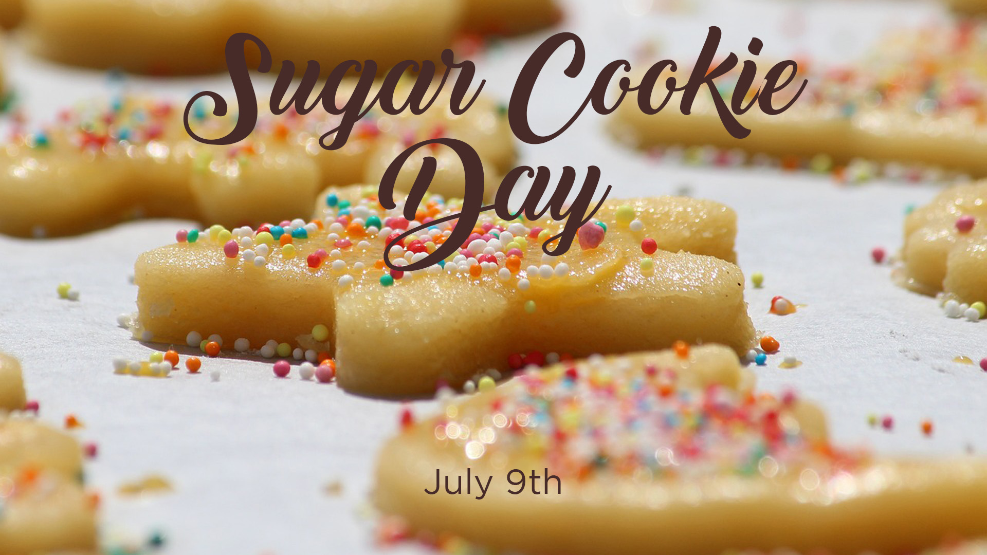 A photo of unbaked sugar cookies laying on a white paper and have a abundant amount of colorful sprinkles. The one cookie that is in the front center of the image is mainly in focus. Along the top of the graphic there is brown cursive font that reads Sugar Cookie Day. Toward the bottom of the graphic a brown font reads July 9th.