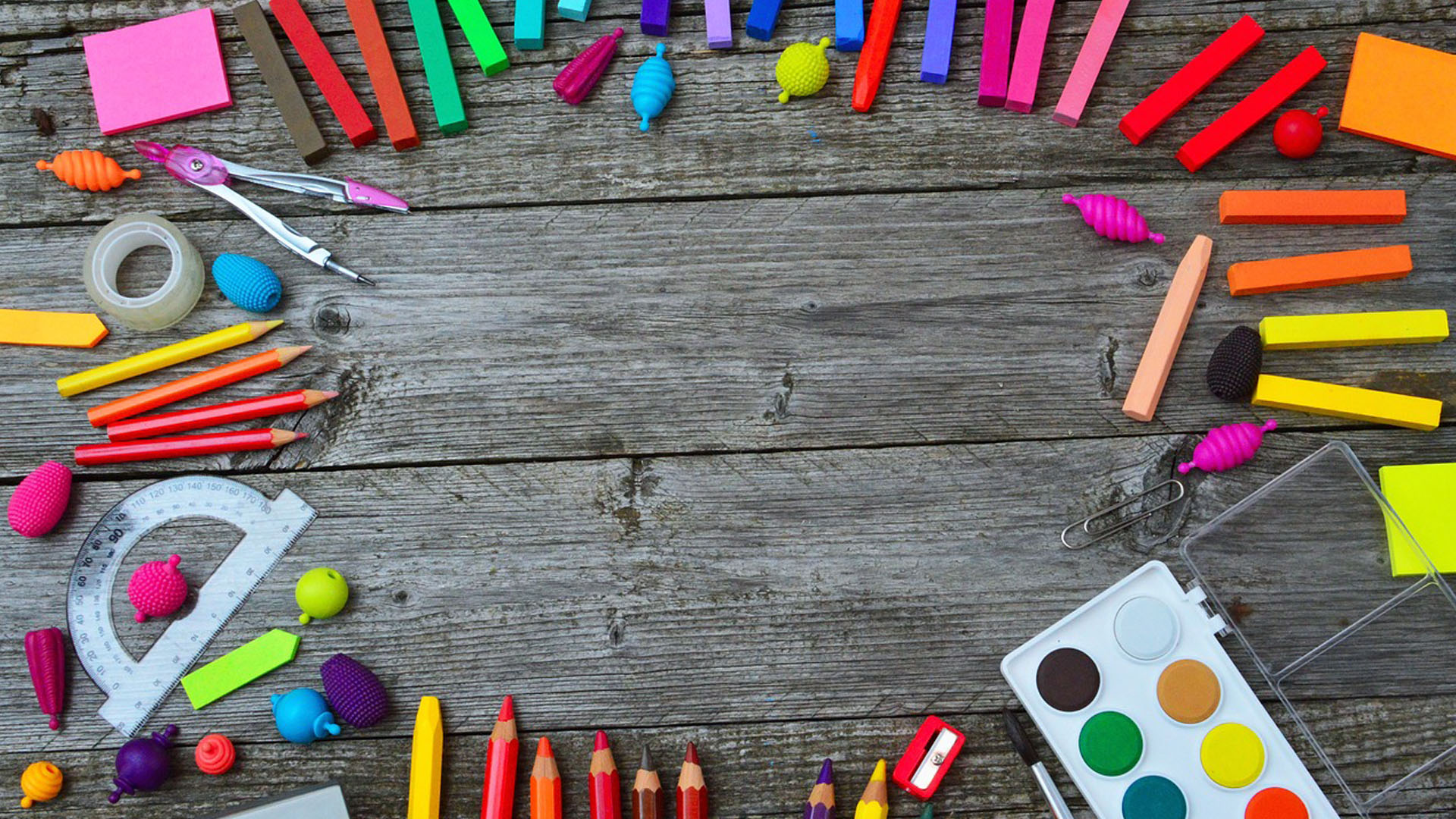 a photo of different colored school supplies ringing around the edges of the image. All of these items are sitting on a wooden porch. Some of the different items include: oil pastels, colored pencils, a paperclips, a compas, a few sticky notes.