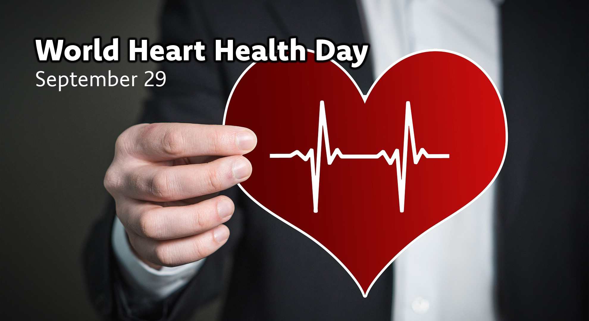 A photos of a person in a black suite and white shirt holds a red heart with a white cardiogram line on it. At the top left in white sans serif letters reads World Heart Health Day September 29