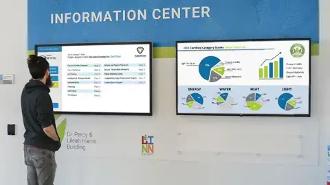 A guy standing and viewing an informational wall. The wall has graphic panels in blue and green and two large screens hanging. On the screens are matrix number for the building and circle graphic charts.