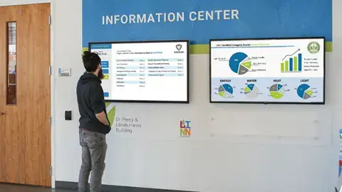 A guy standing and viewing an informational wall. The wall has graphic panels in blue and green and two large screens hanging. On the screens are matrix number for the building and circle graphic charts.