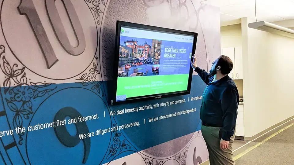 A hallway wall with custom designed wallpaper mural of a clock and a blue stripe with the company mission runs across. Hanging on the wall is a touch screen display in bright blue and green to highlight whats going on and resources. An employee is standing in front of the digital sign reading the content.