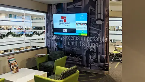 Inside a visual lobby with green chairs and divider wall with graphic wallpaper. A large digital screen hangs on the wall with company news and information.