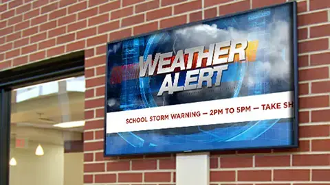 A digital signage screen is mounted to a brick wall. On the screen is a weather alert with a blue background and scrolling information about the alert.