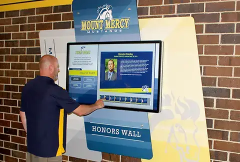 A custom shaped display hangs on a brick wall outside the gym. Gold and blue graphic panels showcase the logo and Honors wall. A large 55" touchscreen TV lets visitors search through the years of athletic recepients