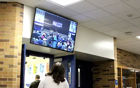 A digital signage screen about the door way entrance to the auditorium runs a live feed of the school event. A student is looking up a the screen to see what is happening