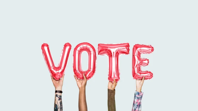 A photo of four hands reaching up from the bottom edge, and holding one red letter balloon each. The balloons spell out the word ‘vote’. The background is a solid gray color.