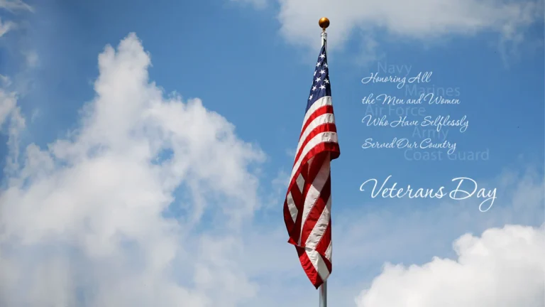 A photo of the sky with fluffy white clouds. The top of a flagpole sits just to the right of the center of the image. An American flag is attached to it. The right of the flag transparent sans and solid serif words are stacked alternately on top of each other. First the transparent words read ‘Navy, Marines, Air Force, Army, Coast Guard’. The serif words are ‘Honoring All, the Men and Women, Who Have Selflessly, Served Our Country, Veterans Day’. All the words are white in color.