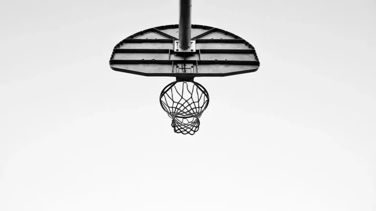 A black and white photo of a basketball hoop. The photo is taken from directly below the net, and is looking up towards an empty sky.