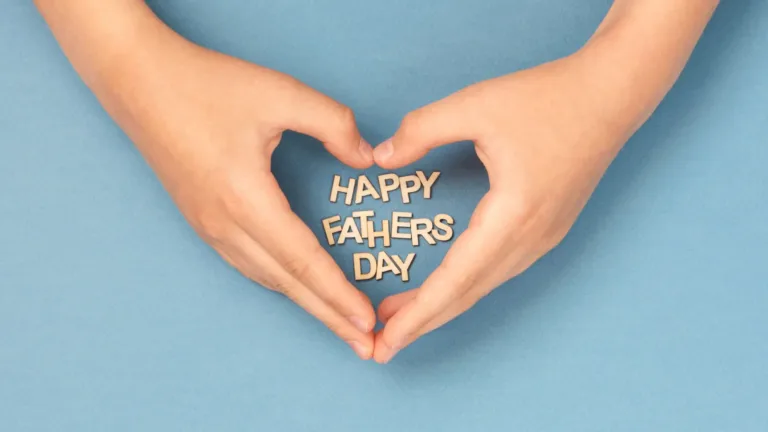 Two hands stretch into the photo from the top edge. The hands are making a heart shape in the center. Inside the heart says ‘Happy Fathers Day’ in small, wooden letters. All on top of a solid white blue background.