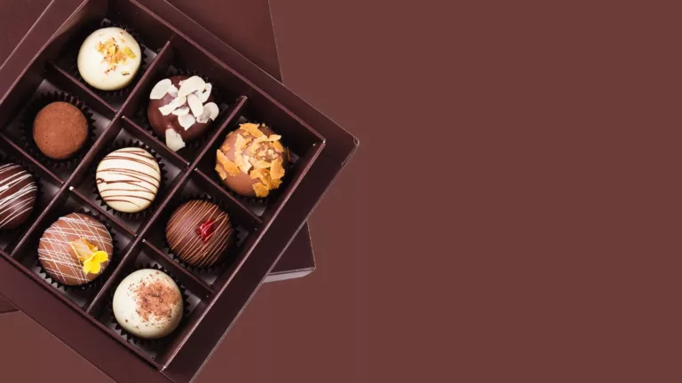 A birds eye photo of a brown square box of nine chocolate candies. The box is tilted and on the left side of the graphic. Its top and left corners are cut off by the graphics edges. The box sits on a solid brown background.