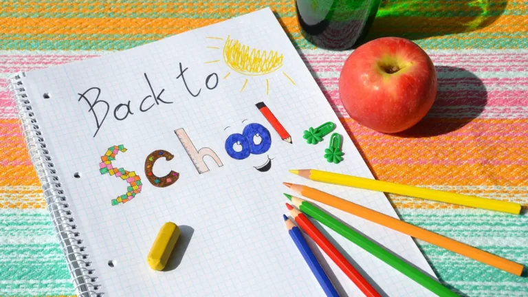 A photo of a tabletop that is covered in a rainbow striped tablecloth. A spiral bound notebook lays open. ‘Back to School’ is written in the notebook. ‘Back to’ is written in pencil. ‘School’ is written and colored in with colored pencils. A small yellow eraser, five color pencils, and two green paper clips also sit on top of the notebook. A red apple and the bottom of a green plastic reusable water bottle are to the top right of the notebook.