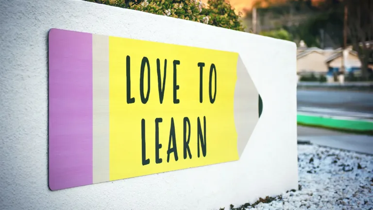 A photo of a cement retaining wall near a road blurred out in the background. On the wall is a large cut out paper in the shape of a pencil. It is also decorated to look like a yellow pencil with a pink eraser. Written on the pencil in bold, black, all-caps letter is ‘LOVE TO LEARN.’