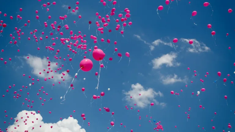 A photo of hundreds of pink balloons floating upwards into the blue sky with fluffy white clouds.