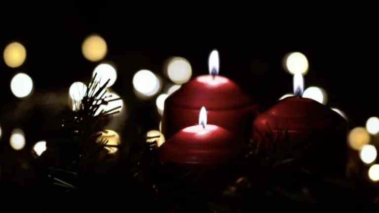 A video of three red pillar candles surrounded by a wreath. The room is quite dark and is illuminated by the candles and some out of focus lights that are scattered circles in the background.