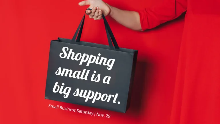 A solid red background. A person standing and wearing a red, long-sleeved dress is partly seen against the right edge. Their right arm is stretched out along the tog edge and is holding a black, rectangular gift bag that hangs into the center of the graphic. On top of the bag are bold,white, cursive, sans letters reading, “Shopping small is a big support.” In small white sans letters below the bag say, “Small Business Saturday | Nov. 29.”