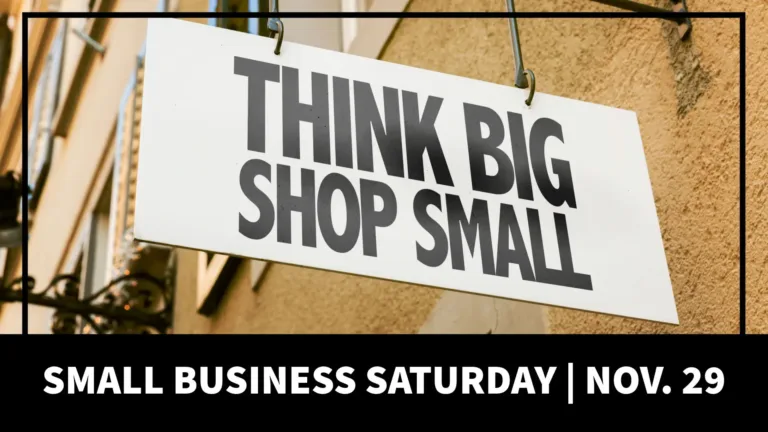 A photo of a white sign hanging off of a tan brick building. It reads, in bold, black sans letters, “THINK BIG SHOP SMALL.” A thin black line border is inset along the top and side edges. The bottom of the graphic is covered in a thick, black rectangle. Inside it reads, “SMALL BUSINESS SATURDAY | NOV. 29” in bold, white sans letters.