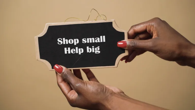 A still thumbnail for a video. A tan background with two hands holding a small chalkboard sign stretch from the bottom and right edge into the video center. This signs says, "Shop small Help big," in white sans letters.