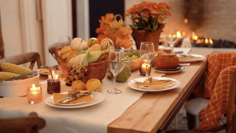 A still thumbnail for a video. A set wooden dining table in front of a fireplace. Fall baskets of leaves, gourds, corn, and potatoes are placed in the center. Candles, wine glasses, plates, and a white runner are also on the table.