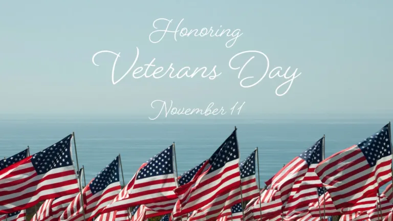 A photo of rows of American flags against a foggy blue sea skyline. The flags cover the bottom edge, while the sky and sea fill the rest of the photo. Stacked, in the center, above the flags are thin, white, cursive words reading, “Honoring Veterans Day November 11.”