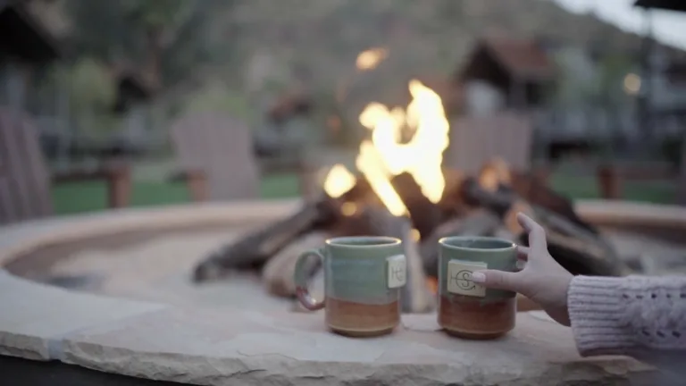 A video of a campfire out of focus with wooden lawn chairs, a large hill, and wooden playground in the background. In focus are wit teal and brown ceramic mugs sitting on the firepit’s stone edge. A hand reaches into the video from the right edge and grabs the nearest mug. It takes the mug off screen back off the right edge.
