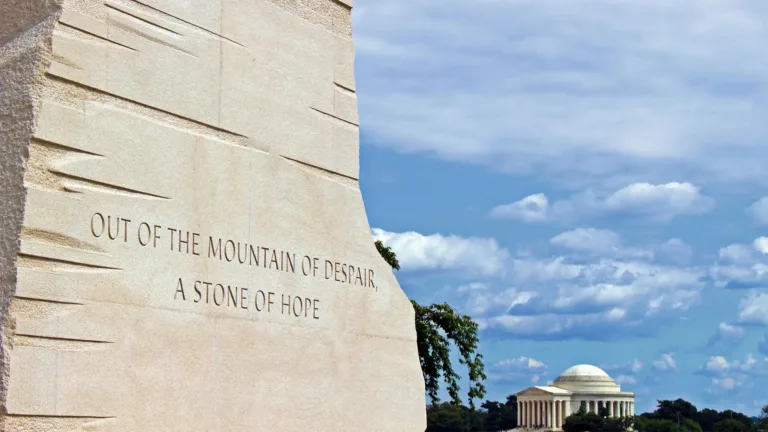 A photo of the part of the Martin Luther King Jr. monument located in Washington D.C. The part shown appears to the monument’s side. All that is visible on the stone are the etched words, “OUT OF THE MOUNTAIN OF DISPAIR, A STONE OF HOPE.” The shown part of the monument takes up the left side of the image. The right side shows the Lincoln Memorial off in the distance against a bright blue sky with fluffy white clouds.