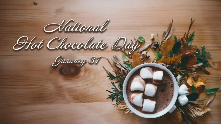 A birds-eye view of a white mug full of hot chocolate and marshmallows on top of a wooden table. The mug is located in the bottom right cinder and has dried leaves around it. In white cursive letters near the top left corner and center say, “National Hot Chocolate Day January 31.” The words have a small drop shadow.