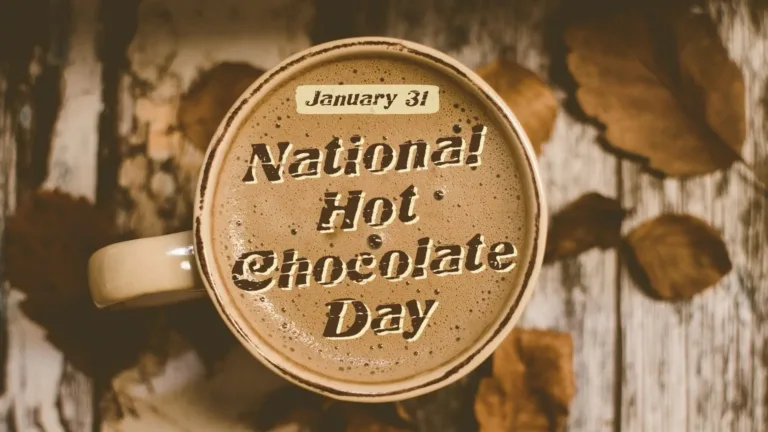 A birds-eye view of a tan mug full of hot chocolate on top of a rustic, white painted, wooden table with dried leaves around it. Overtop the mug and hot chocolate are the stacked words, "National Hot Chocolate Day.” A distressed, brown, serif font with a small off-white drop shadow was used. Over top to words is a small off-white rectangle with date January 31 written in small brown serif letters.