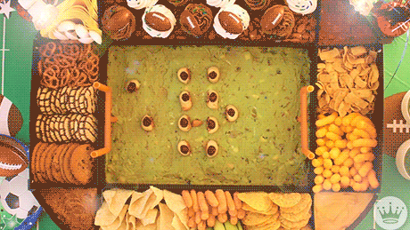 A birds-eye view of a tray of food in the shape of a rectangle football stadium. The tray sits on a green table top with different sports graphics on it. The center of the tray is filled with guacamole with a orange football goal post on either end. Their are small pigs-in-a-blanket sausages moving around the guac field. Their football flies up, covers the gif, then falls back down revealing the words, 'Let's Watch Football' in bold white letters has replaced the players on the field. The stands of the outer tray sections are filled with jittering crackers, cookies, dips, and veggies.