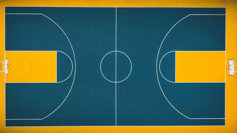 A birds-eye photo of a basketball court. The courts free throw lanes, and sidelines are yellow. The rest of the court is dark blue with white lines. The backboards are also white.