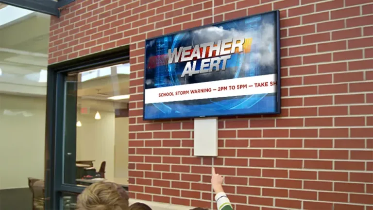 A digital sign on a brick wall in a school plays a weather alert graphic to inform students in the hall of the alert