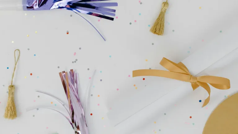 A birds-eye photo of a white tabletop covered in small multi colored confetti, two gold tassels, a rolled up diploma with a gold ribbon, and a pink and blue noise makers.
