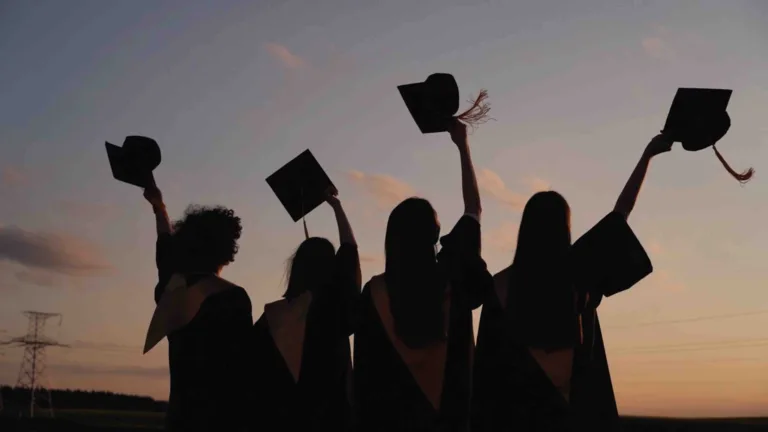 Four people stand next to each other and wave their graduation caps up in the air. They are back lit against a sunset.