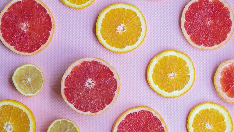 A light pink table cloth covers the birds-eye view of the photo. On top of the table top are circle slices of citrus fruits, almost creating diagonal lines. There are pink grapefruits, oranges and a few lemon slices.