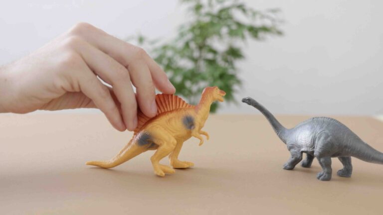 A wooden table top with a black plastic brachiosaurus toy on top of it near the right edge. A hand walks in a yellow plastic spinosaurus toy from the left edge and to the brachiosaurus.