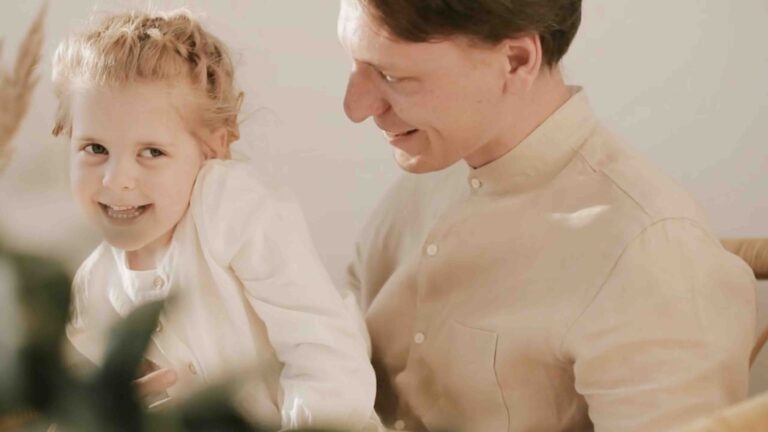 A video of a father with his young daughter sitting on his lap. They are smiling, laughing, and hugging each other.