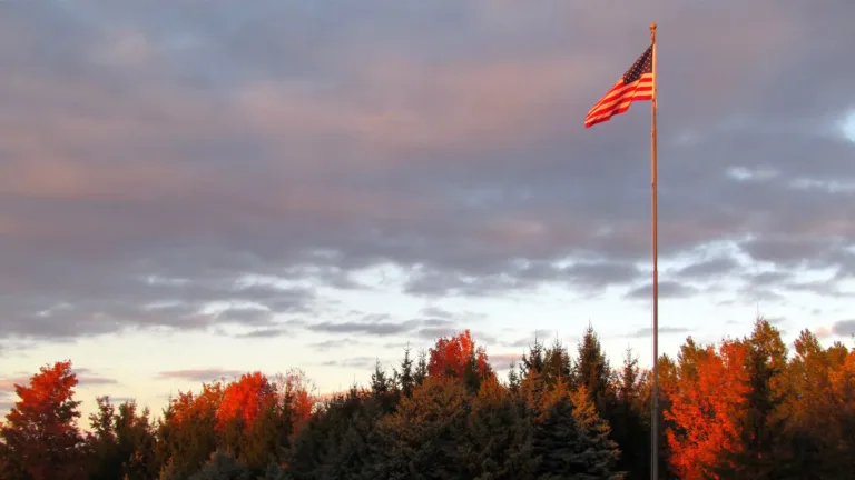 A photograph of the American Flag in a pole against a cloudy blue sky and pine trees long the bottom edge. The flag pole is located near the right edge. The photo appears to have been taken during the sunset.