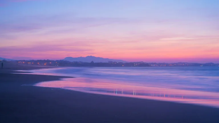 A photo of the pink sunset, close to dusk, over the a beach. The darkness makes the sand almost black and the sea is reflecting the sunset. Faint city lights can be seen off in the distance.