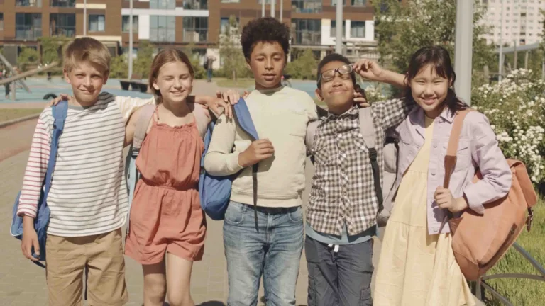 Five kids standing outside with their arms around each other. The kids seem happy and all have backpacks on.