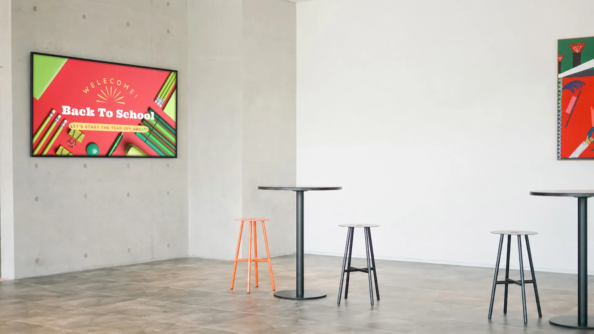 Feature image: A industrial room with cement floors and walls. There are a couple of black bar height circle tables and stools in the room. In the edge of back wall a red painting can be seen just before the rest of it gets cut off the right side. In the adjacent wall on the left side is a tv screen with a back to school free monthly graphic by Arreya.